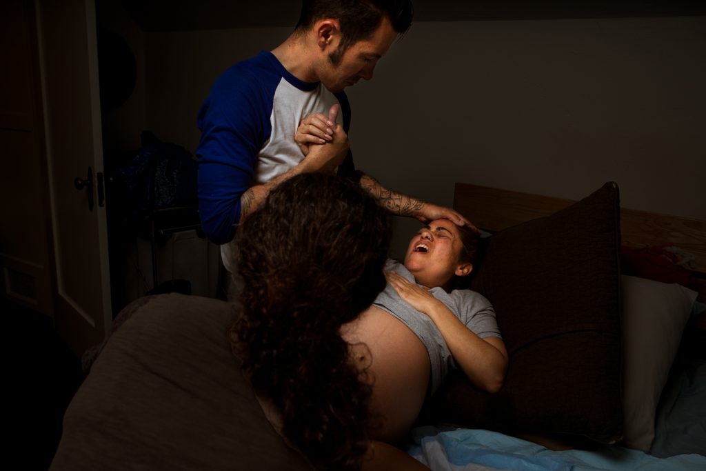 Birth photo of a laboring mother holding onto her partner while midwife delivers the baby.