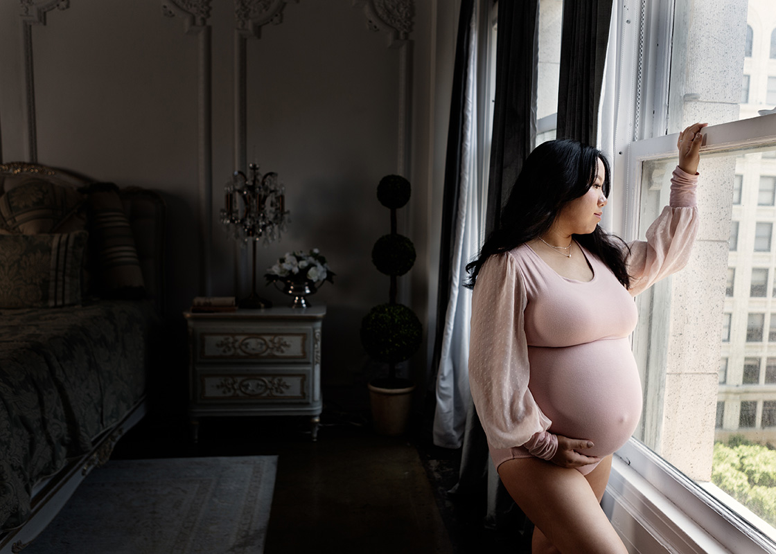 Color maternity portrait of a pregnant woman gazing out a bedroom window in a pink body suit