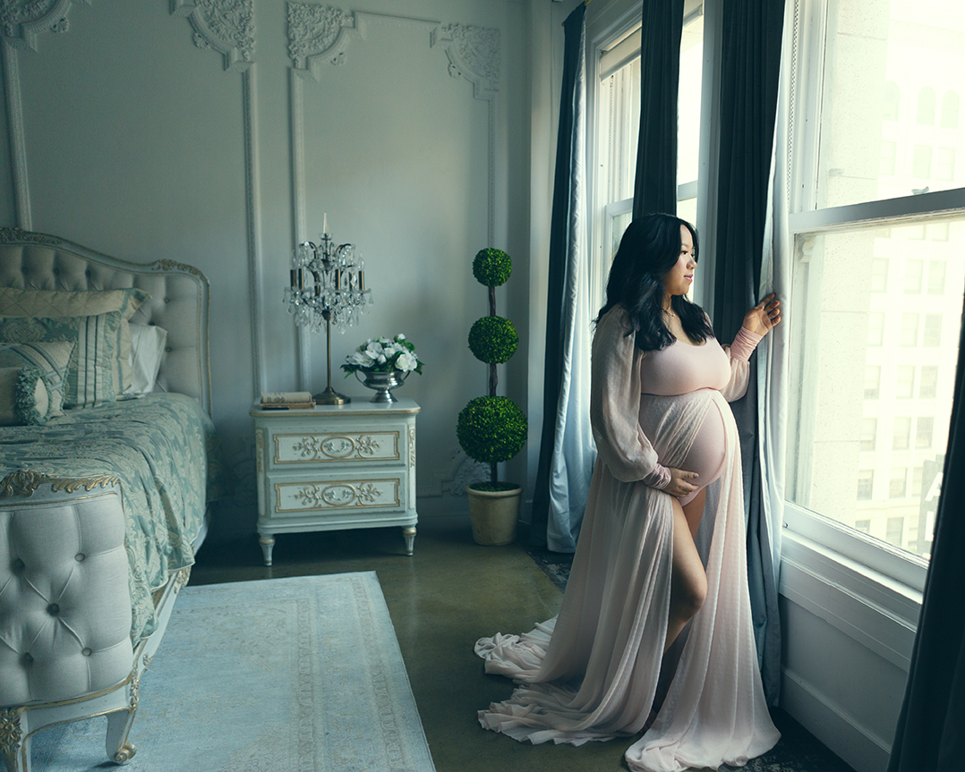 Pregnancy photo of an asian woman in a pink maternity gown looking out of a window