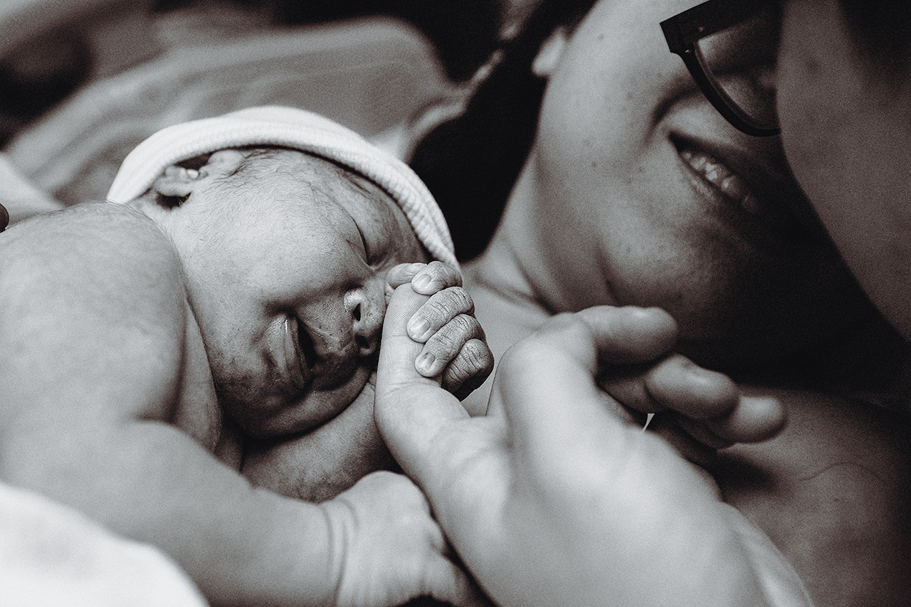 black and white birth photo of a mom holding her baby, smiling and dad kisses her and holds baby's hand.