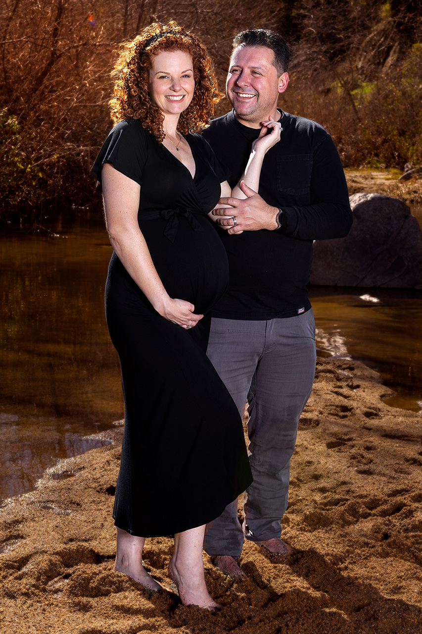 Outdoor maternity photo of a woman and her husband smiling. She is in a black dress and holding her belly. Photo by Leona Darnell