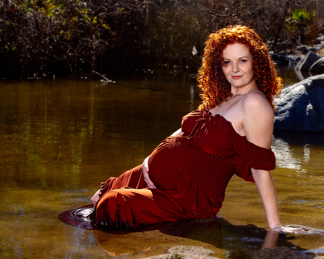outdoor maternity photoshoot showing a pregnant woman in a brown dress sitting in a creek by Leona Darnell