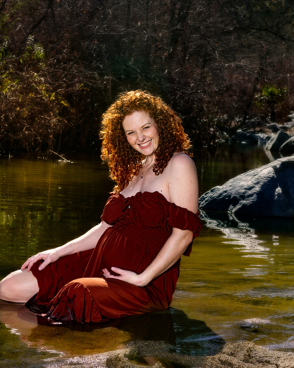 outside maternity photo of a pregnant woman smiling and sitting in a creek by Leona Darnell