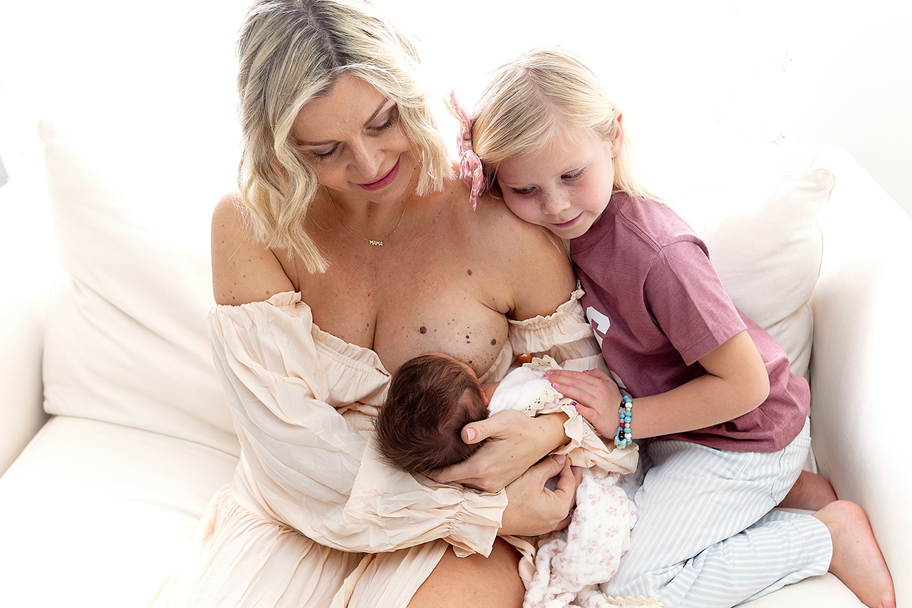 Lifestyle newborn photo session of a mother nursing her newborn and a sibling sister watching by Birth and Beauty.