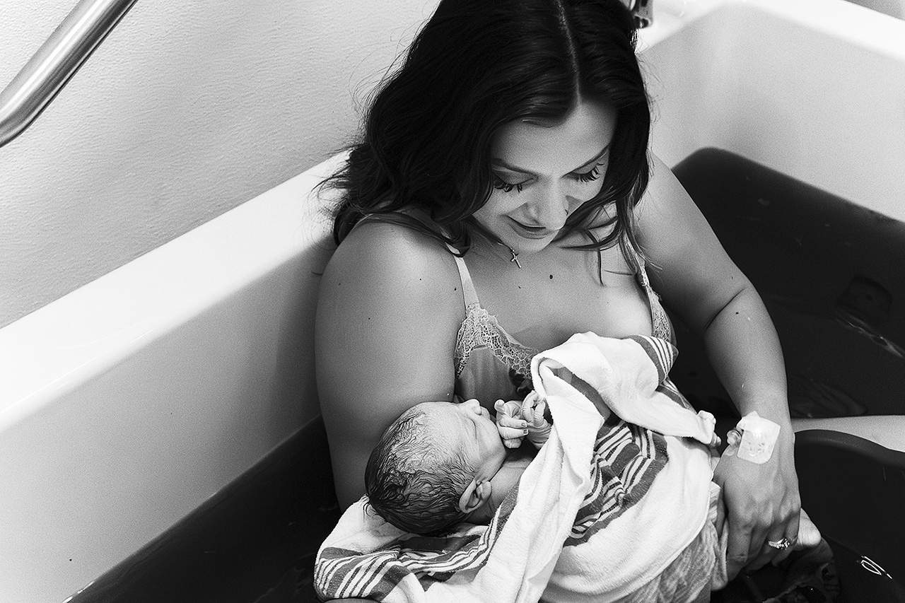 Black and white birth photo of a new mom in a birth center tub holding her daughter.