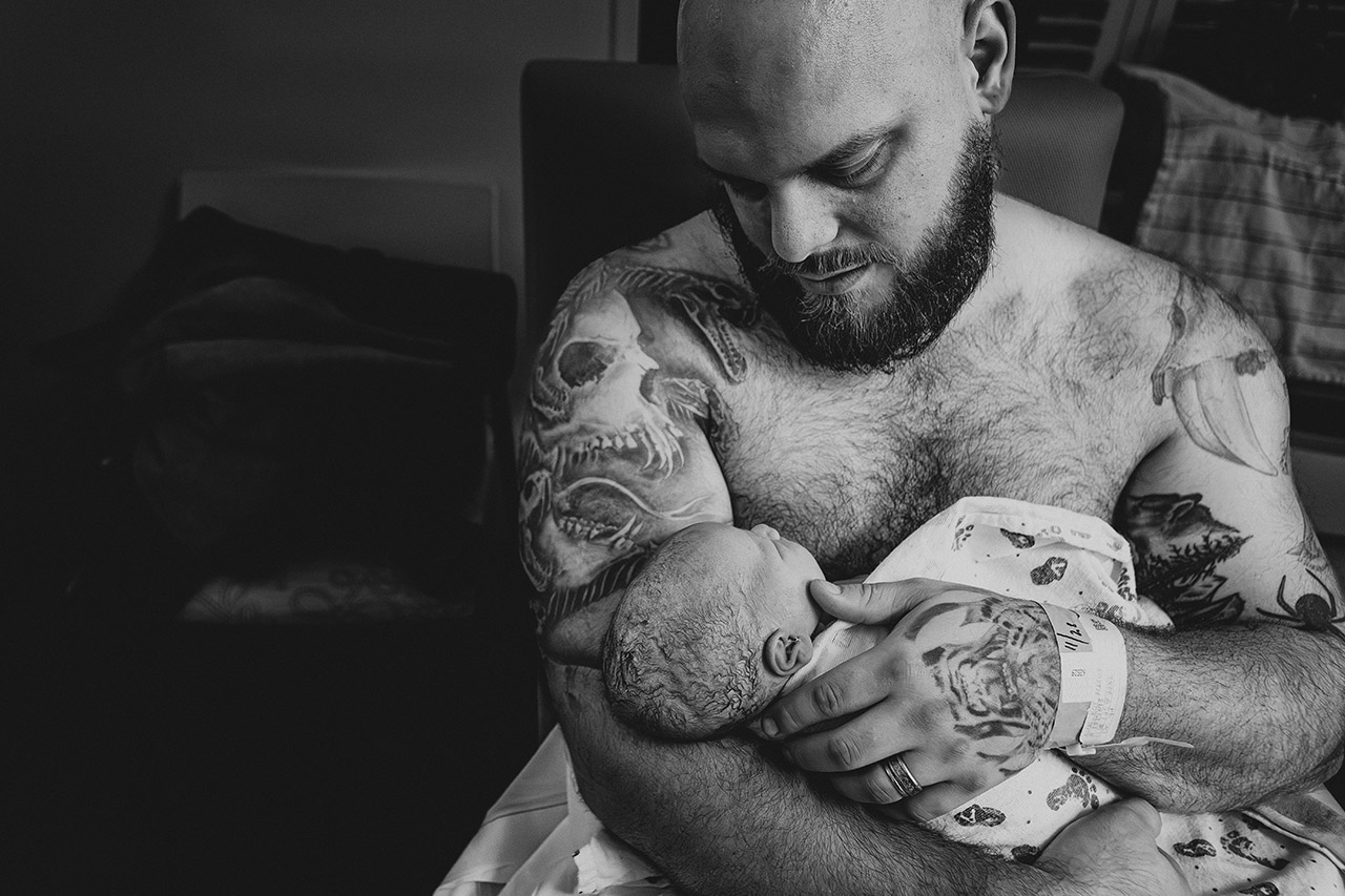 Black and white image of a father holding his new baby against his chest in skin-to-skin contact, photographed by Leona Darnell.