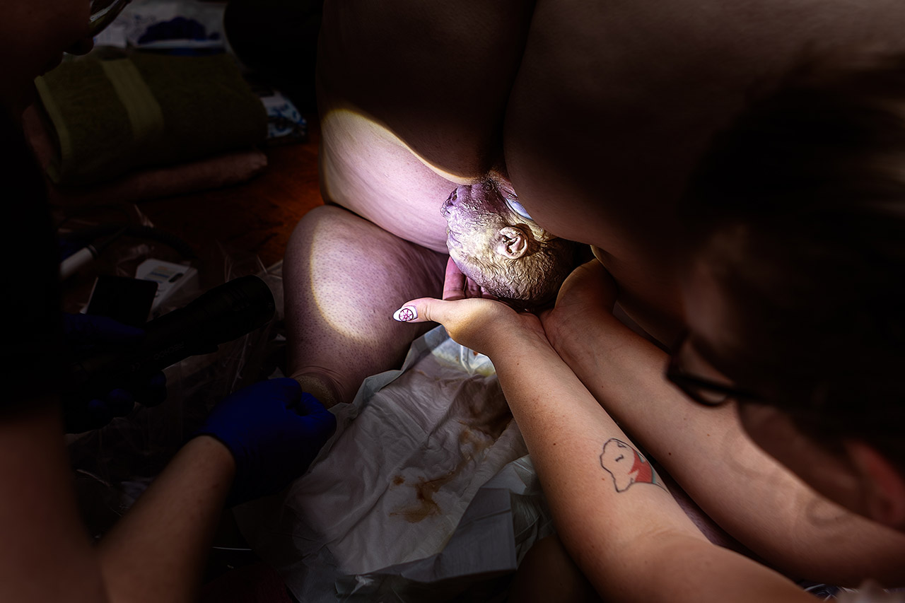 Color birth photography capturing a tender moment as a baby is welcomed into the world, with a family member’s hands waiting nearby in loving anticipation. Photo by Leona Darnell.