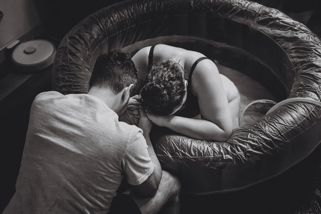 Black and white birth photo of a woman laboring in a birth pool with her husband. Photographed by Leona Darnell.