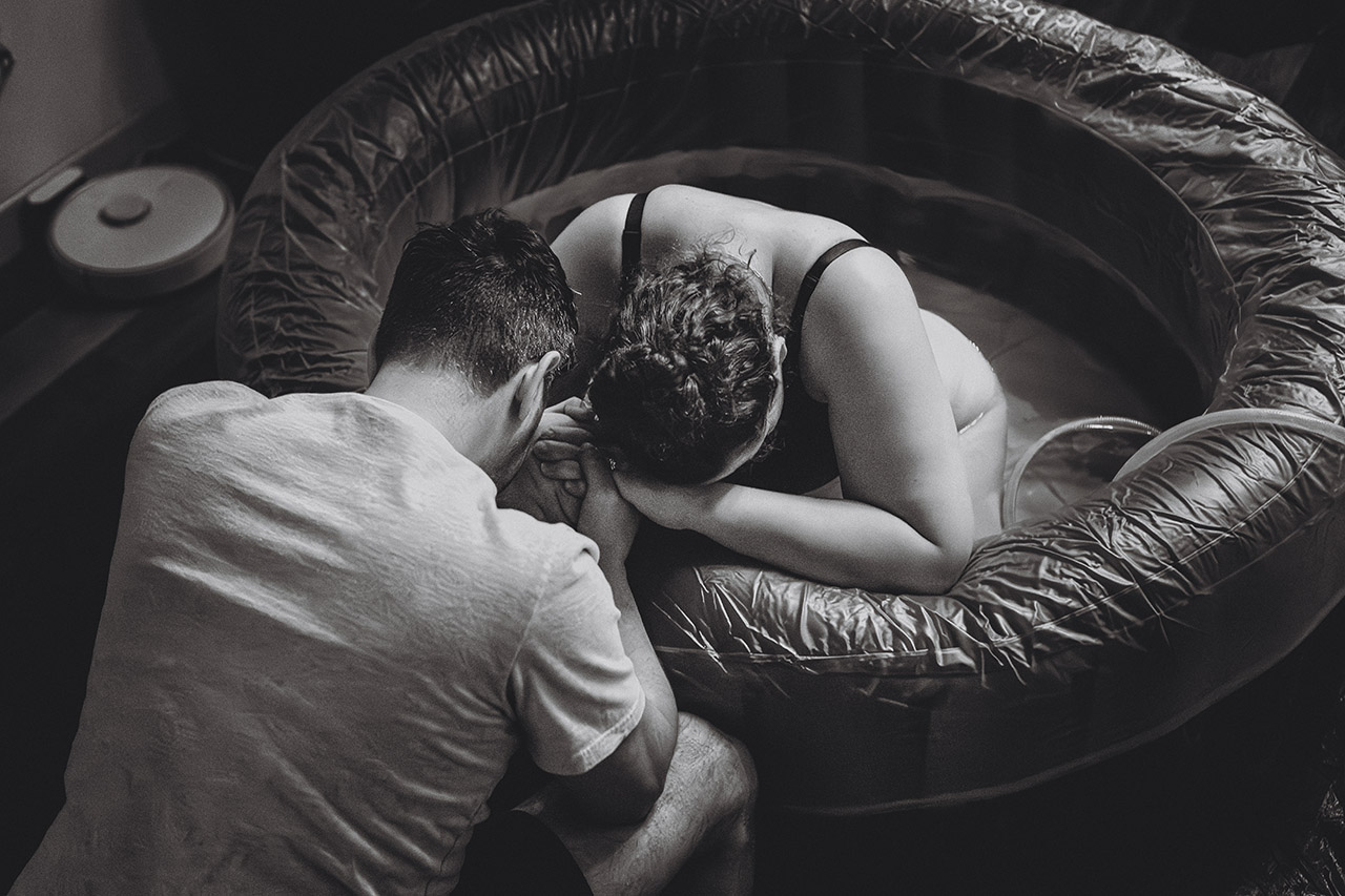Black and white birth photo of a woman laboring in a birth pool with her husband. Photographed by Leona Darnell.