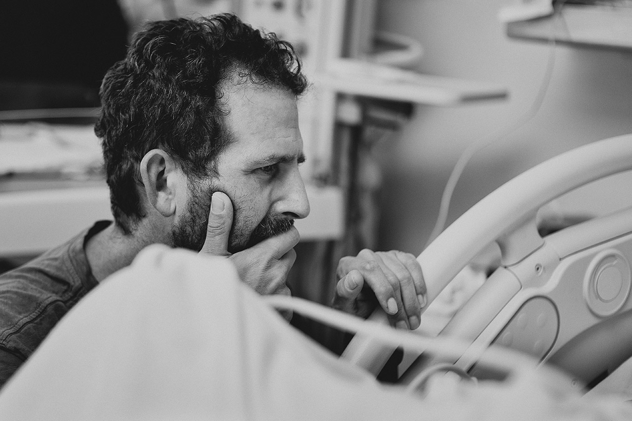 An attentive father holds his partner’s hand during labor in a Los Angeles hospital, staying close and focused as he supports her through the birth of their fifth baby. Leona Darnell captures the quiet connection between them.