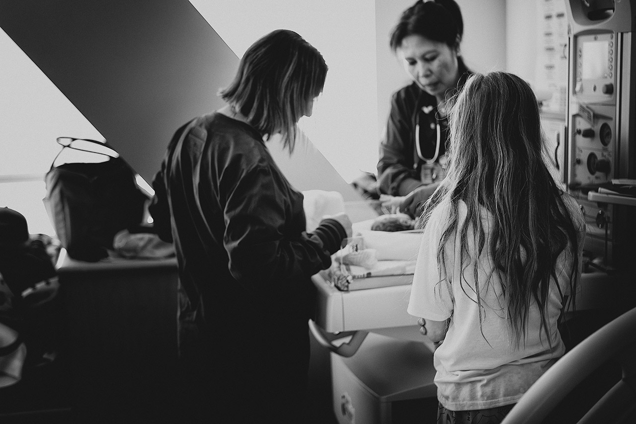A black and white birth image of the oldest daughter watches her newborn sister being examined. Birth documentation by Leona Darnell. 