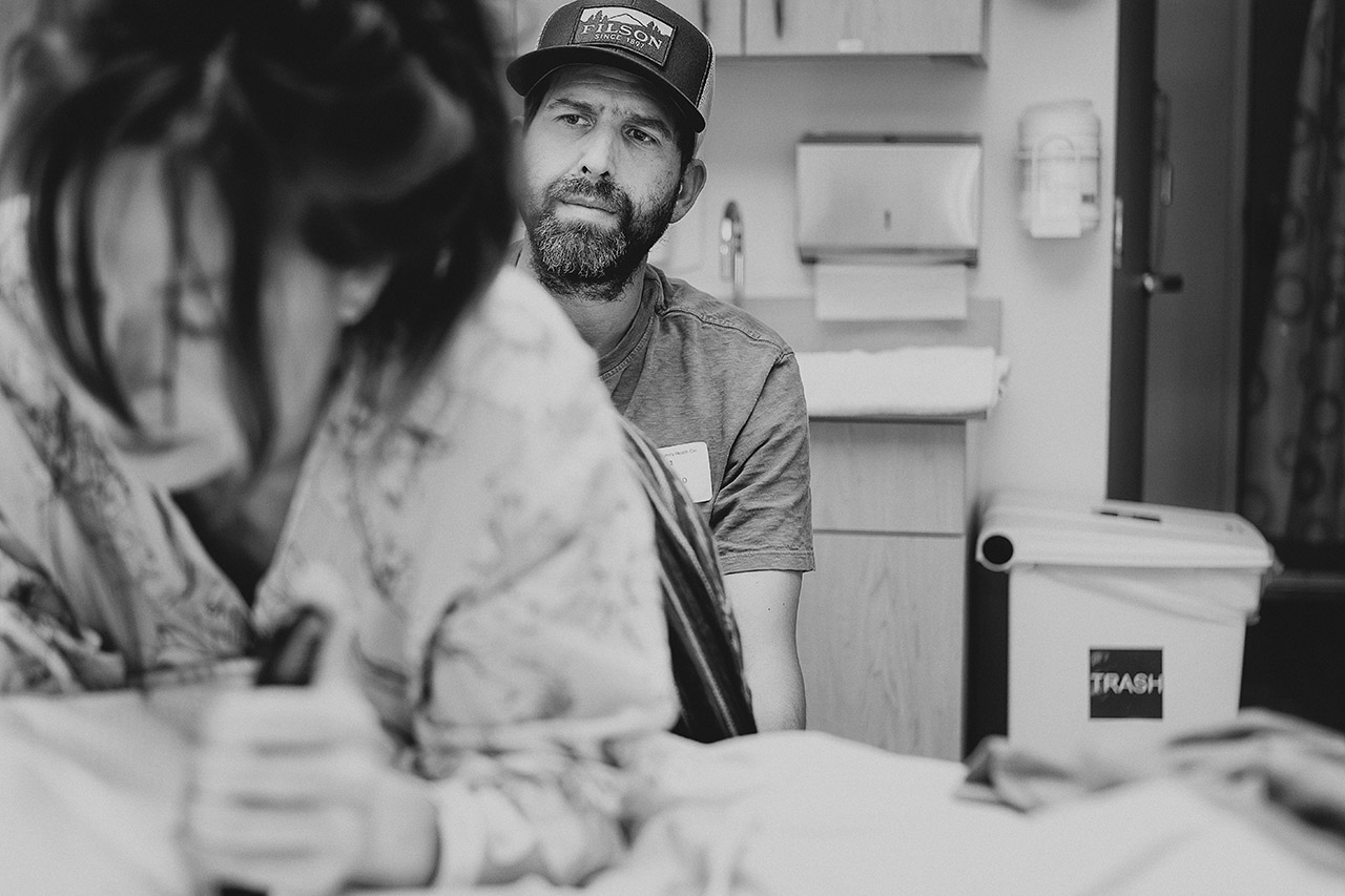 A father looks lovingly toward his partner during labor in a Los Angeles hospital, remaining calm and attentive as she prepares to welcome their fifth baby. Photographed by Leona Darnell.